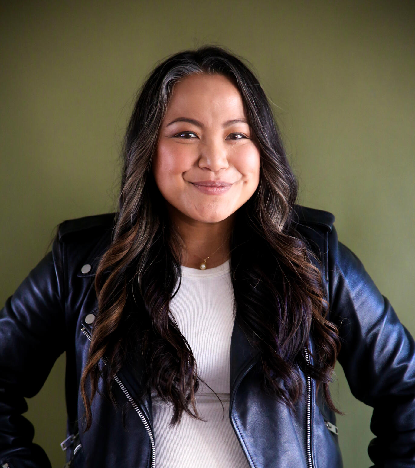 Kat Evasco Asian woman with long wavy dark brown hair smiling and wearing a dark leather jacket and white shirt