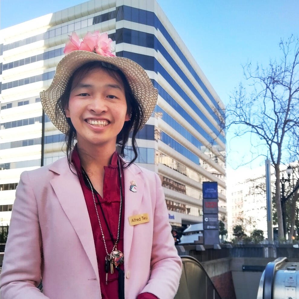 Alfred Twu Person with long black hair and medium light skin tone wears a red colored shirt, pink jacket, and wide-brimmed hat with pink flowers on it, standing in front of BART subway entrance