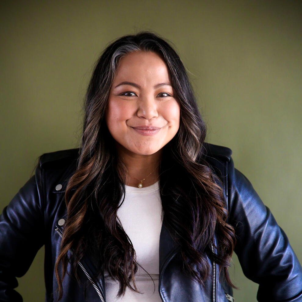Kat Evasco Asian woman with long wavy dark brown hair smiling and wearing a dark leather jacket and white shirt