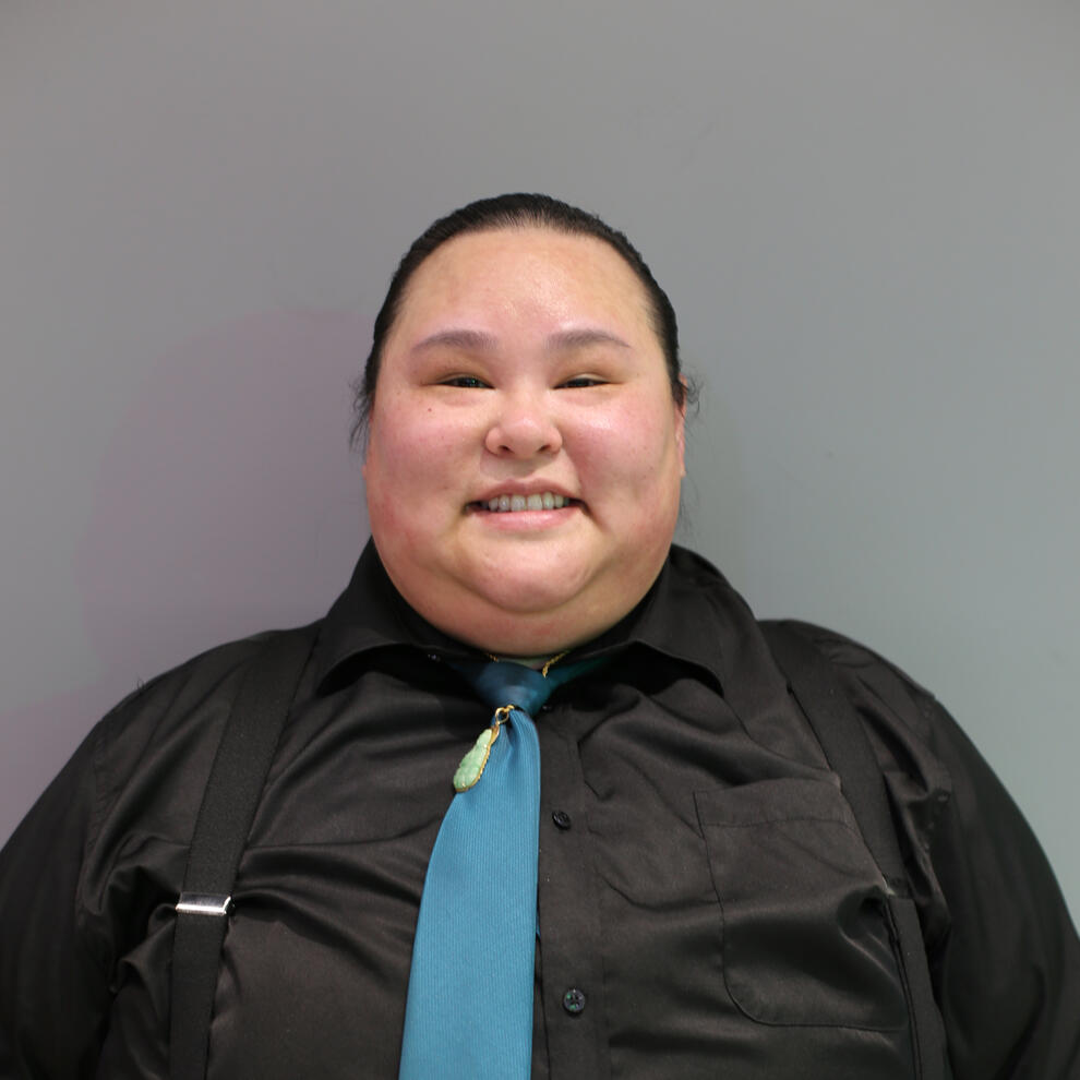 Total Nguyen Person with medium skin and dark hair that is tied back smiles at camera wearing a dark collared shirt, teal tie, and black suspenders.